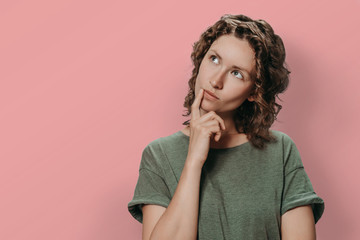 Fototapeta na wymiar Young female with curly hair keeping her finger on her lips while thinking about question, dreaming, trying to make up her mind or solve the problem. Girl being deep in thoughts, Isolated on pink. 
