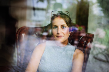 Young woman with glasses sits in a leather armchair behind a window and looks into the camera