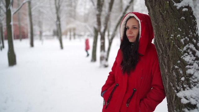 Portrait of young woman in red jacket and hood in winter season. Beautiful lady stands near tree and looks away. Fluffy snow envelops everything around.