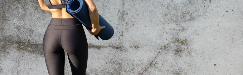 Fototapeten Ein sportliches schlankes Mädchen in Leggings und einem Top steht mit einer Trainingsmatte in der Nähe einer Betonwand und ruht sich zwischen den Übungen aus. © Mountains Hunter