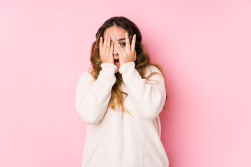 Young curvy woman posing in a pink background isolated blink through fingers frightened and nervous.