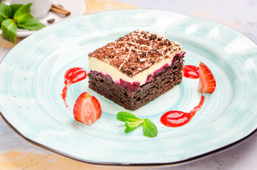 Delicious piece of chocolate cake with sour cream, cherry impregnation, mint, strawberry on a blue plate, close-up on a background. Homemade dessert with cream and cherry