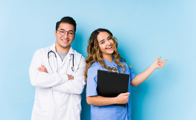 Young doctor couple posing in a blue background isolated excited pointing with forefingers away.