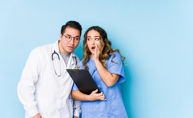 Young doctor couple posing in a blue background isolated is saying a secret hot braking news and looking aside