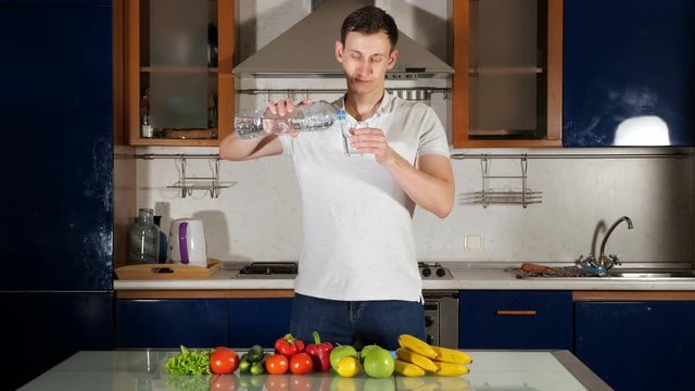 tall young man pours water into glass from bottle and drinks assuaging thirst and standing at table with vegetables