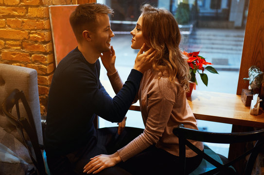 young man and woman tenderly hugges and touches each other in cozy cafe in front of big window.
