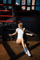 Naklejka premium Beautiful blonde in white bodysuit posing at camera. A woman sits on a hardwood floor near a boxing ring