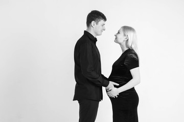 Pregnant woman and her husband hugging and holding onto your stomach in black clothes on a white background. Black and white picture. Beautiful married couple. Copy, empty space for text