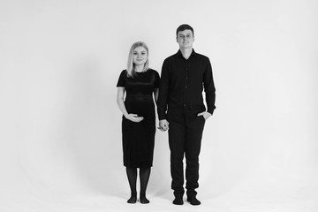 Pregnant woman and her husband holding hands in black clothes on a white background. Black and white picture. Beautiful married couple. Copy, empty space for text