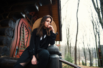 Beautiful girl with long hair and straw hat sitting on the railing of an old wooden house and looks into the camera. Autumn walks on a cloudy day in the forest