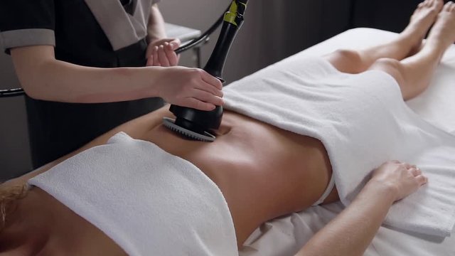 Unknown experienced cosmetologist which holding special massage instrument doing anti cellulite procedure on woman's belly