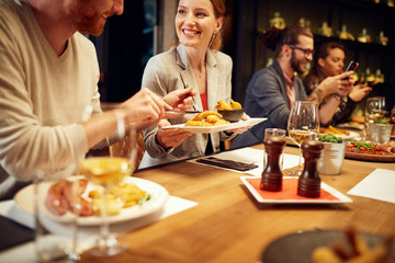 Handsome caucasian ginger taking food out of his girlfriend's plate while sitting in restaurant for dinner. In background are their friends.