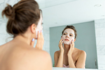 Young woman applying cosmetic mask on face while looking at reflection in mirror.