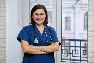 Smiling adult woman doctor in blue uniform stethoscope with folded arms, confident female medic looking at camera, standing near the window in clinic