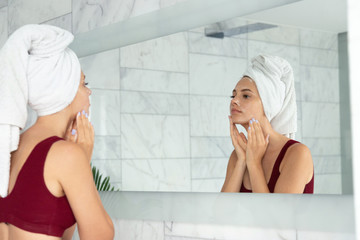 Mirror reflection of beautiful young woman wearing bath towel on head with face cream on skin.