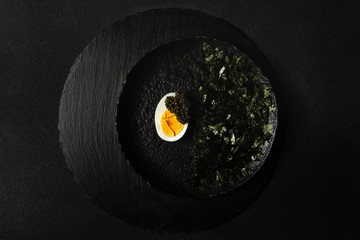 Appetizer of sturgeon caviar, half of boiled egg, shredded nori on black plate on black background