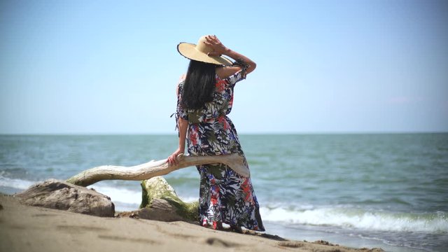 Romantic woman sitting alone is watching the sea view on overturned dry tree trunk on beach. Girl in hat looking view landscape. Female dressed in colorful long skirt dress.Emotional alone sentimental