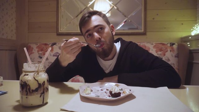 A bearded guy neatly with a spoon eats a delicious cheesecake in a restaurant.