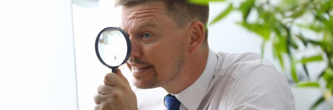 Portrait of businessman in office at computer. Manager in classic shirt and tie looking at pc monitor through magnifier attentively. Blurred background