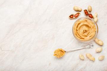 Peanut butter in glass plate with peanuts in shell, peeled peanuts, vintage spoon with peanut butter. Creamy peanut paste flat lay with place for text on white marble background for cooking breakfast.
