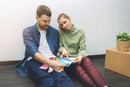 Young Couple Sitting On The Floor In The Room And Choosing Paint Color From Swatch For New Interior Design