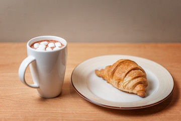 A croissant on a white plate next to a mug of cocoa and marshmallows on a wooden table.