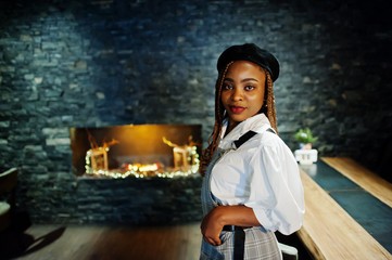 Obraz premium African american woman in overalls and beret in room with wooden table and fireplace.