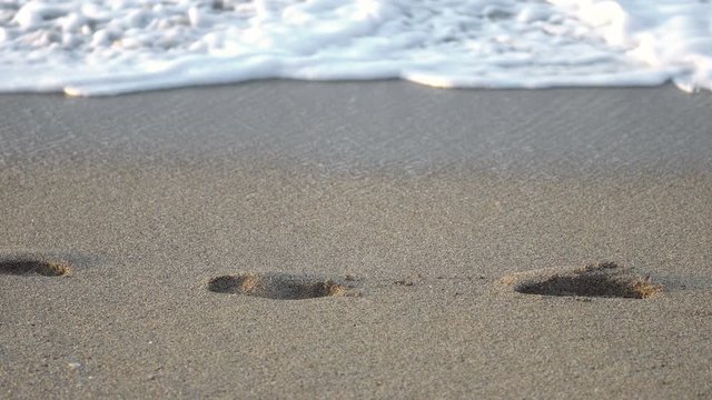 Woman's bare feet walk on the sand on the beach and leave footprints and waves roll on the shore
