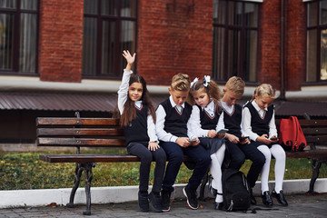 Group of kids in school uniform sits on the bench outdoors together near education building