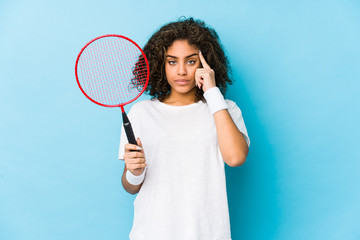 Young african american woman playing badminton pointing temple with finger, thinking, focused on a task.