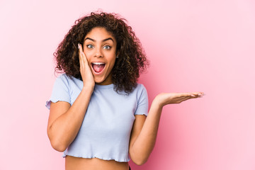 Young african american woman against a pink background holds copy space on a palm, keep hand over cheek. Amazed and delighted.
