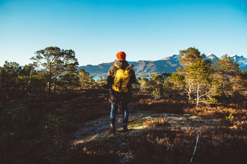 Traveler man is standing in the middle of a forest with a yellow backpack and guitar on background of mountains and lake. Wearing a in a red hat. Place for text or advertising. Shoot from the back