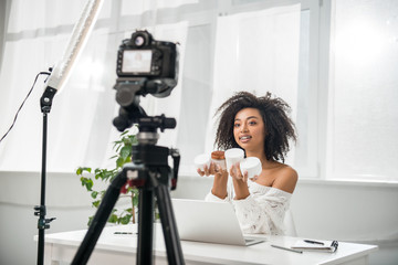 selective focus of attractive african american influencer in braces holding containers with cosmetic cream near digital camera