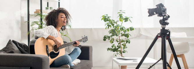 panoramic shot of beautiful african american influencer in braces playing acoustic guitar near digital camera