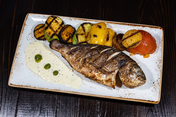 Grilled dorado fish with vegetables on a white plate on a dark wooden background