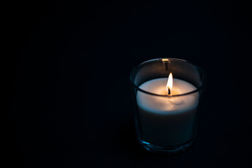 Burning white candle in transparent glass on a black background.
