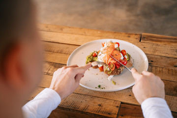 Young man enjoys big and tasty breakfast at cafe. Delicious sandwich or toast with avocado and smoked salmon on top. Healthy food concept.