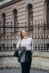 Fototapeta na wymiar Outdoor photo of blonde lady posing on architecture background in autumn day.Close up fashion street style portrait.wearing dark casual trousers,creamy sweater and gray coat or jacket.Fashion concept.