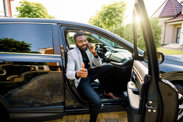 Fototapeta premium Successful handsome African man businessman and boss sitting in the car, talking on the phone, smiling, looking at camera and showing thumb up. Business concept