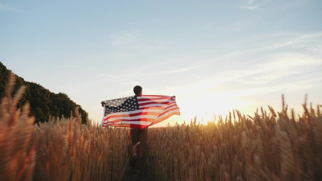 Rear view: Man walking with flag of America in the hands in the wheat field. Slow motion shot