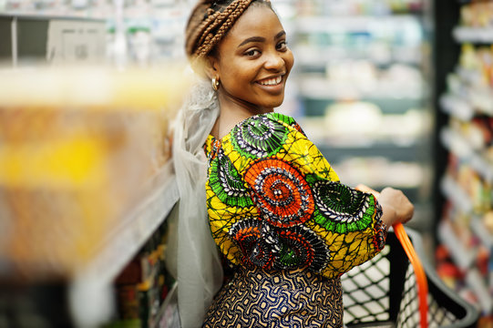 Happy african woman in traditional clothes and veil looking product at grocery store, shopping in supermarket.  Afro black women costumer with basket buying food at the market.