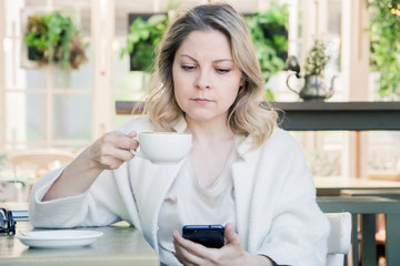 Cute blonde girl sitting in a cafe with a Cup of coffee with a mobile device, spending time on social networks