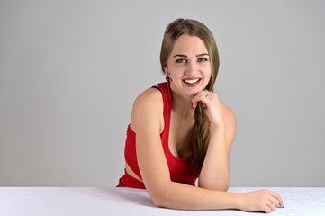 Fototapeta premium Universal concept female portrait on a white background. Horizontal photo of a pretty smiling girl with long hair and great makeup sitting at a white table in the studio.