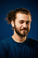 Obraz premium Young charming attractive Caucasian bearded hipster with curly hair dressed in sweater standing in front of blue background and looking at camera.