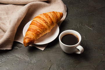 Croissant and cup of coffee with spoon and saucer on a dark background with gray linen cloth