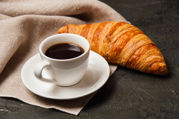 Croissant and cup of coffee and saucer on dark background with a gray linen napkin
