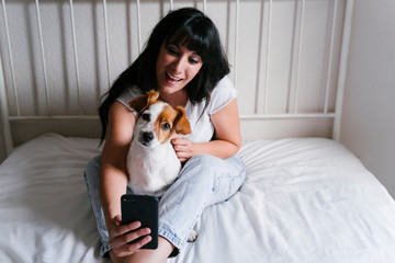 young caucasian woman on bed taking a selfie with mobile phone with her Cute small dog. Love for animals and technology concept. Lifestyle indoors