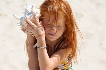 little long-haired red-haired girl with her eyes closed held a large white shell to her ear