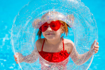 little red-haired girl in a red swimsuit and heart - shaped sunglasses holds a swimming circle in the pool