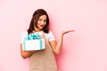 Fototapeta na wymiar Middle age latin woman holding a cake isolated on a pink background showing a copy space on a palm and holding another hand on waist.